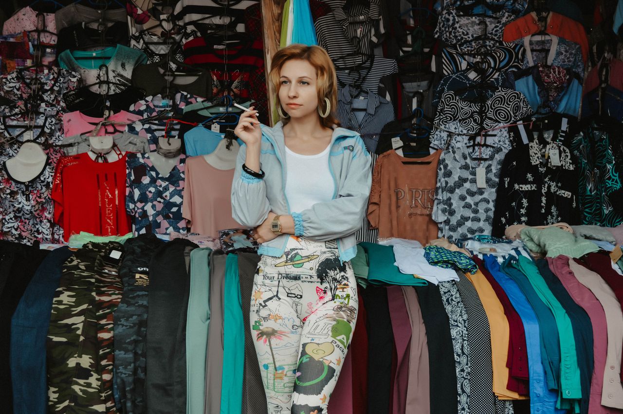 Thoughtful female in casual clothes leaning on shelf with garments while working in market and smoking cigarette